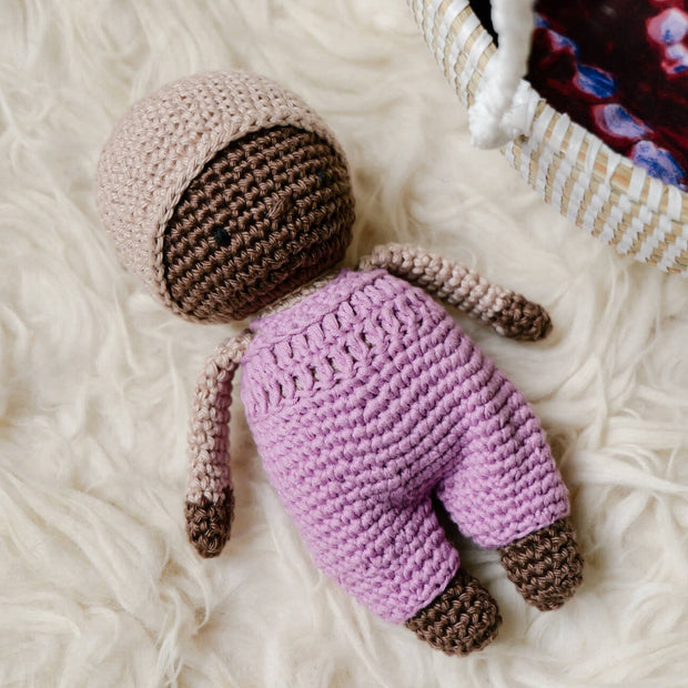 Charming La Petite Senna with a mocha hat and lilac dungarees, showcased with a white basket cot, pillow, mattress, and duvet 