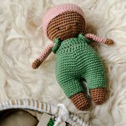 Charming La Petite Senna with a green hat and pink dungarees, showcased with a white basket cot, pillow, mattress, and duvet