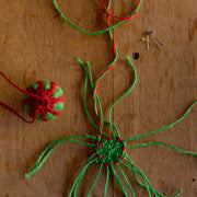 Work in progress a green and red Christmas bauble woven with colourful twine on a wooden table as part of the Weave Your Own Christmas Baubles Kit by la basketry  