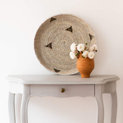 La Basketry woven serving with a triangle pattern laid against a white wall with a bouquet of white flowers