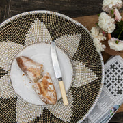 La Basketry woven serving tray with a flower pattern set up on a wooden table with a plate and some bread