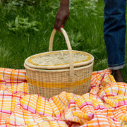 La Basketry picnic basket in natural woven basket with a yellow stripe shown on a tartan blanket about to be lifted by a woman's hand