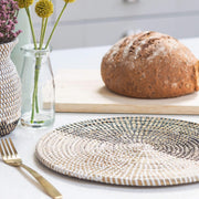a beautiful handwoven african placemat in natural weave with black and white recycled plastic weaved through in half white, half black. by la basketry, shown in a breakfast setting with weaved vase and flowers