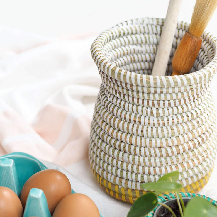 la basketry handwoven vase in white and yellow weave filled with kitchen utensils, shown on a counter with a box of eggs and small basket with a plant in